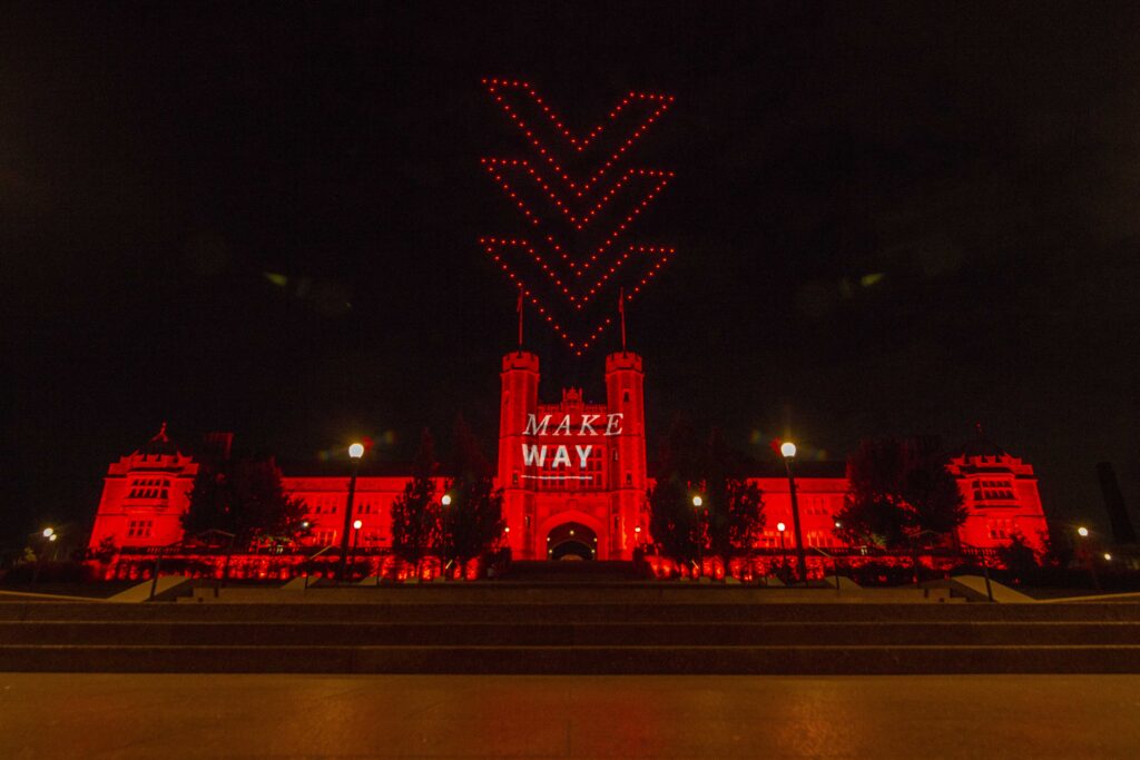 Brookings Hall lit up in red with Make Way logo displayed on building in lights. Red drones above making shape of an arrow toward Brookings