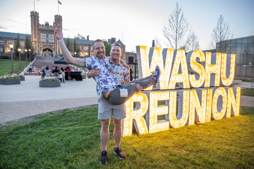 Man holding other man in his arms in front of lit-up WashU Reunion sign
